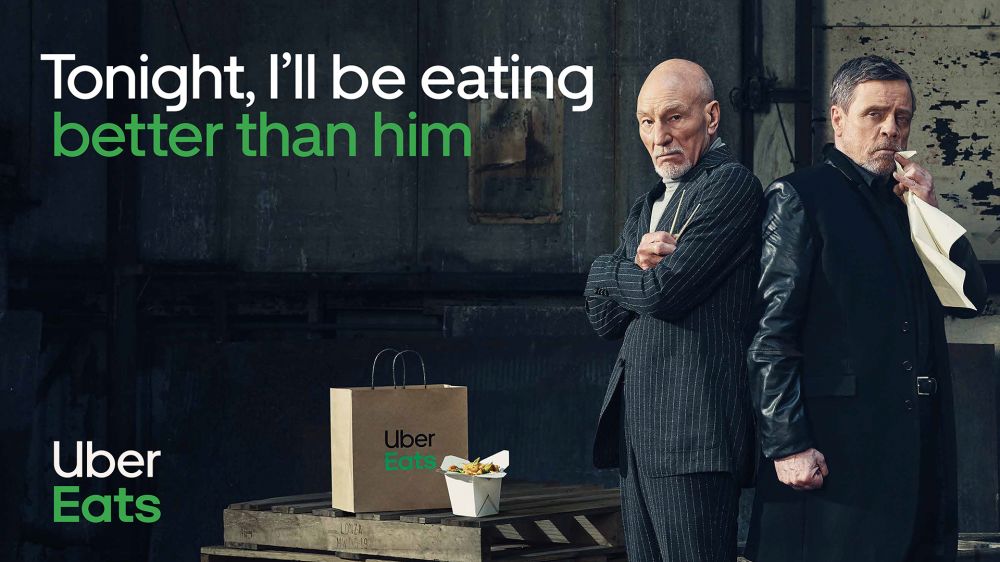Two adult males stand back-to-back with a brown paper Uber Eats bag and a white take-out container in an indoor urban setting.