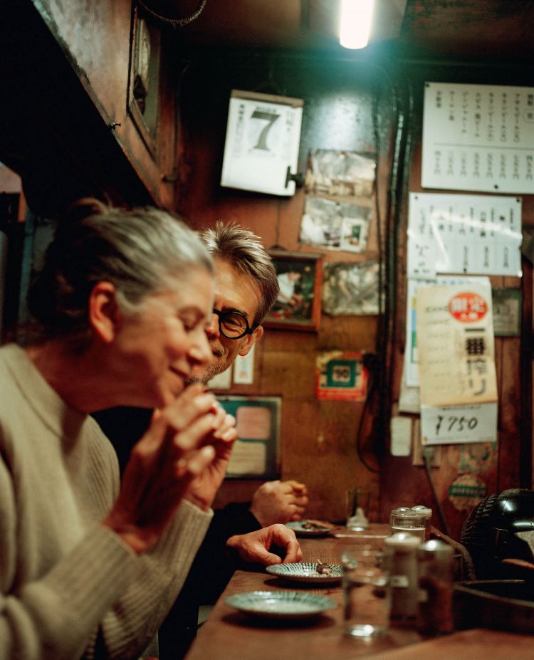Two people at an indoor bar: a woman in a white sweater, a man with glasses, small plates, and various items on the counter.