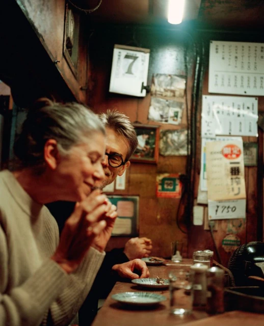 Two people at an indoor bar: a woman in a white sweater, a man with glasses, small plates, and various items on the counter.