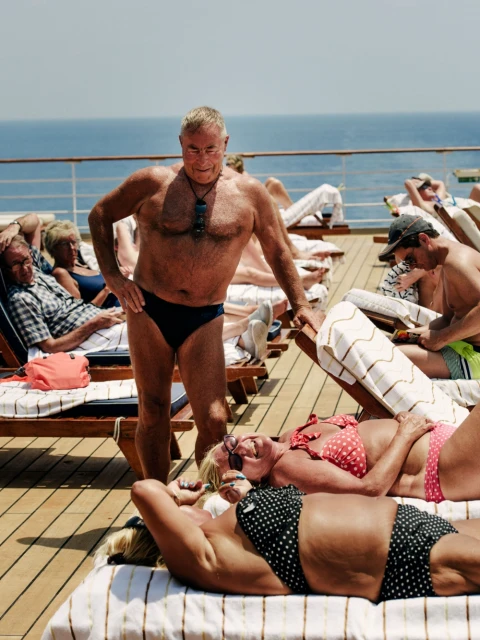 Mature man in swimwear stands on deck with sunbathers, overlooking ocean on cruise ship. Summer travel and leisure concept.