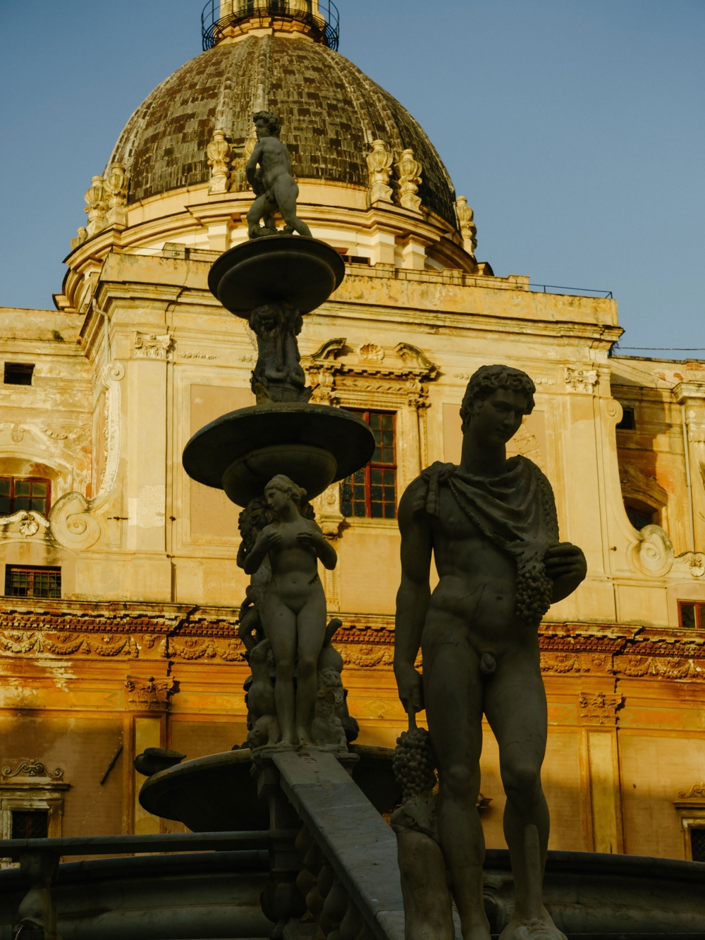 Stone fountain with three nude statues in front of an elaborate building with a dome, bathed in warm light, in an urban setting.