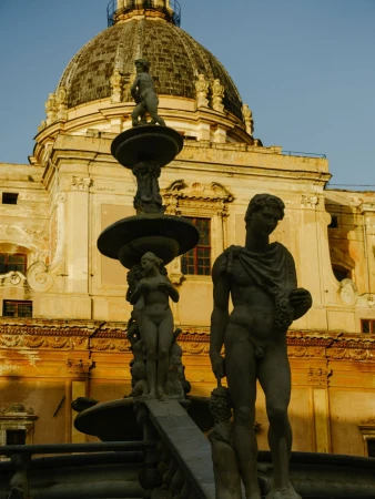 Stone fountain with three nude statues in front of an elaborate building with a dome, bathed in warm light, in an urban setting.