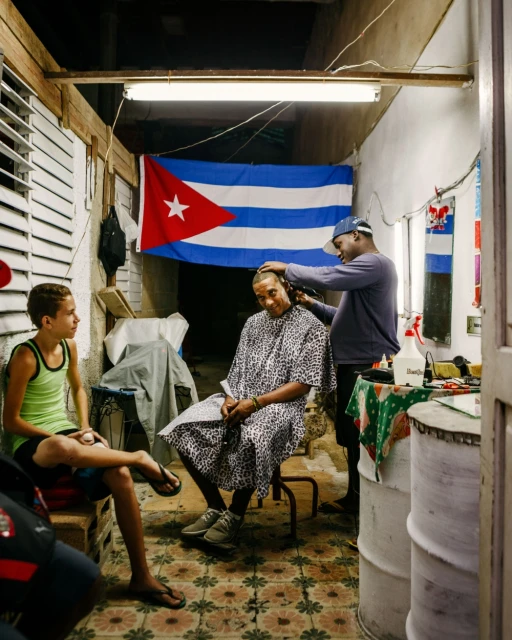 A barber cutting a man's hair inside a barbershop, a boy sits nearby, with a Cuban flag hanging in the background.