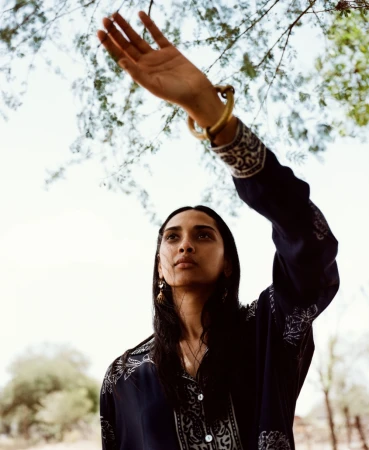Woman in patterned navy tunic raising her hand to shield her face beneath tree branches