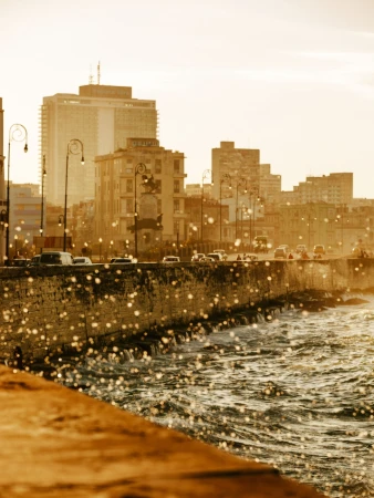 Golden hour view of a coastal sea wall with splashing ocean water, city streetlights, and buildings in the background.