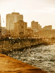 Golden hour view of a coastal sea wall with splashing ocean water, city streetlights, and buildings in the background.