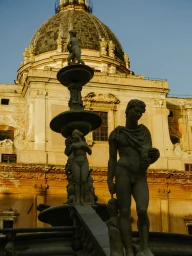 Classical stone statues of figures and a tiered fountain against an ornate historic building with a large dome.