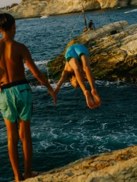 Boy diving into the ocean from rocky cliffs while another boy watches by the water by photographer João Canziani.