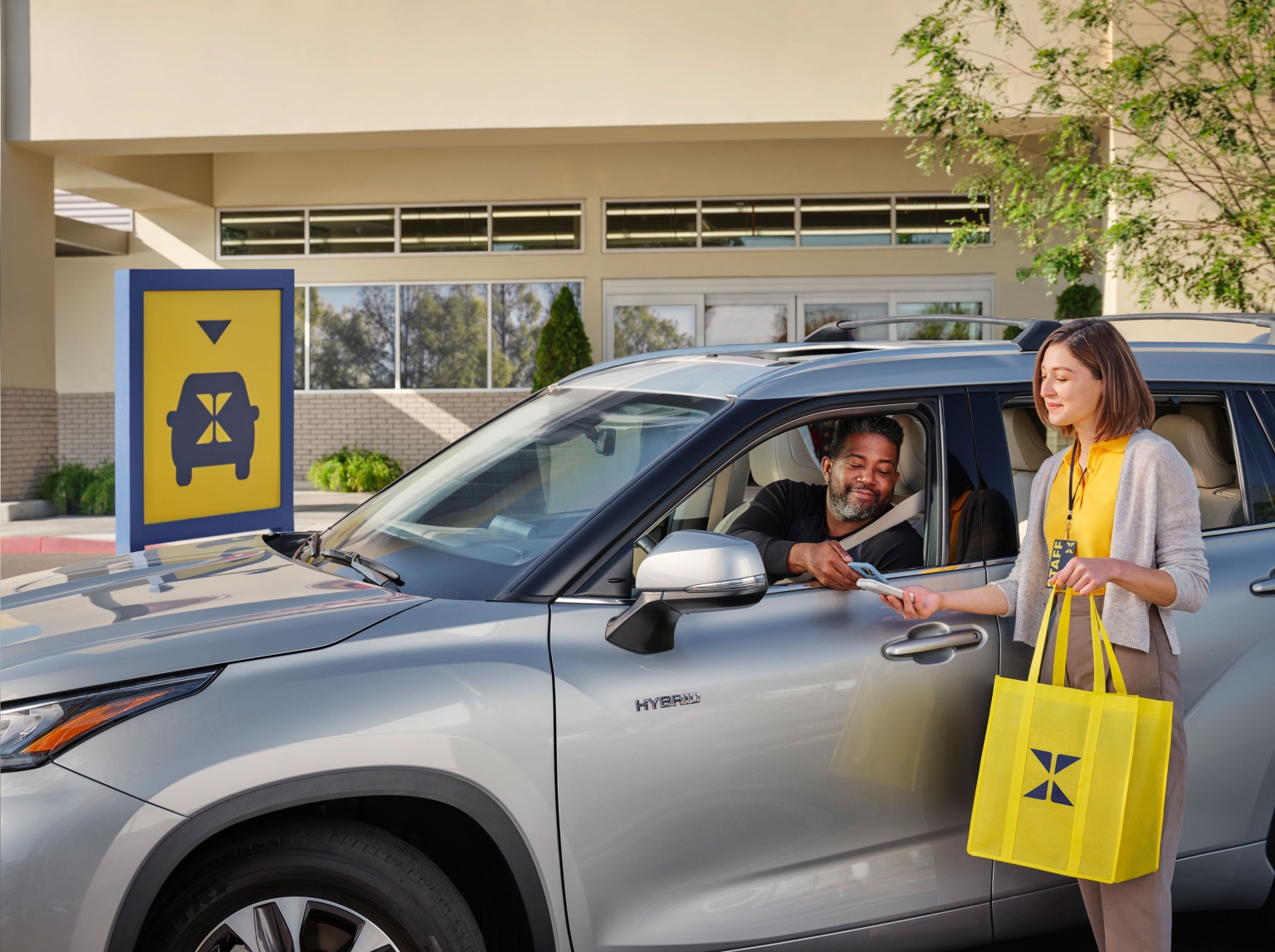 Drive-up pickup service: an adult male in a car receives merchandise from an adult female holding a yellow shopping bag outdoors.
