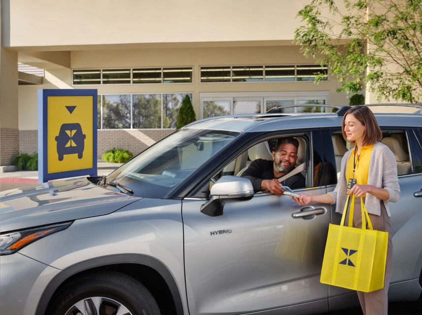 Drive-up pickup service: an adult male in a car receives merchandise from an adult female holding a yellow shopping bag outdoors.