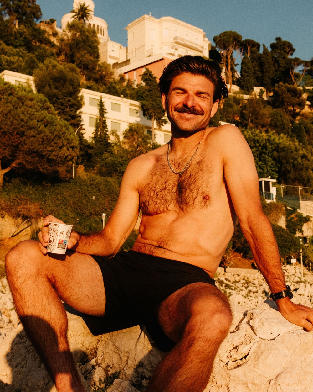 Man with mustache sits shirtless on rocks with beverage. Background: mountains, trees, and buildings on sunny day. Black trunks.