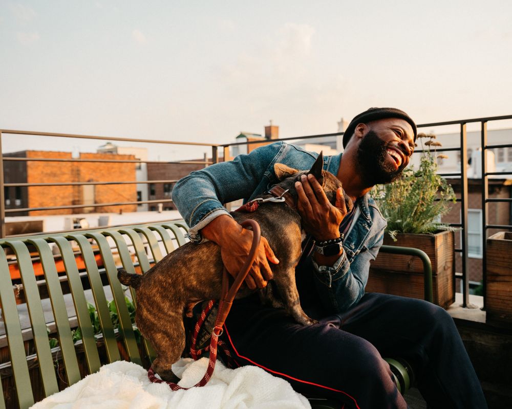 A happy Black man sits on a green bench on a rooftop, hugging his brindle French Bulldog. The man wears a denim jacket and black hat.
