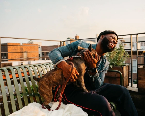 A happy Black man sits on a green bench on a rooftop, hugging his brindle French Bulldog. The man wears a denim jacket and black hat.