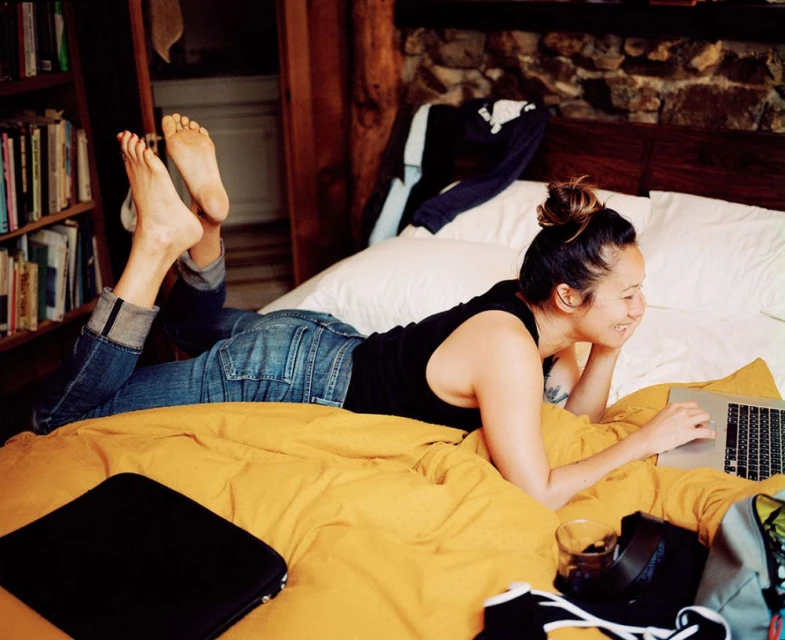 Young woman works on a laptop while lying on a bed with yellow bedding in a casual indoor setting with a bookcase visible.