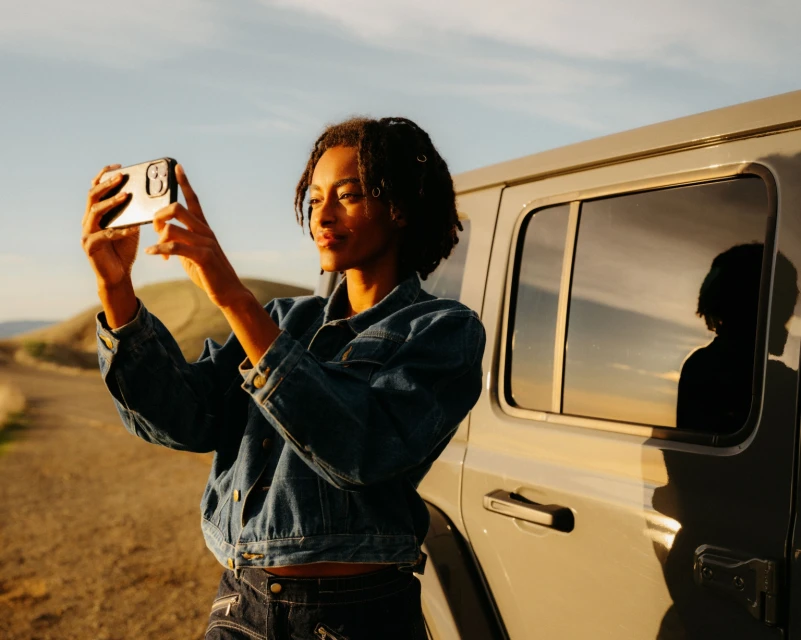 Young Black woman in denim jacket takes a photo with her smartphone next to a silver SUV on a dirt road at sunset.