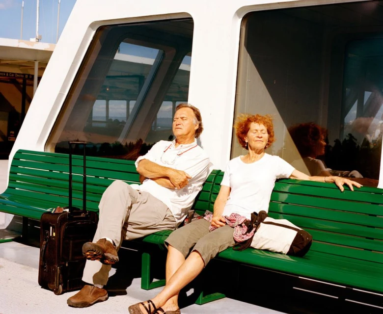 Mature couple relaxing on a green bench outside in sunny weather with luggage near large windows, creating a sense of leisure and travel.