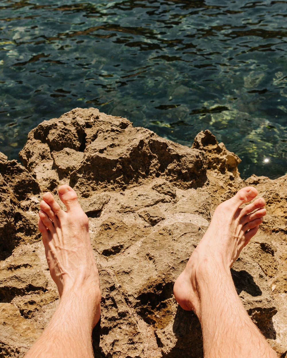 View of a person's feet resting on rocks by clear ocean water on a sunny day, creating a relaxing and natural coastal scene.