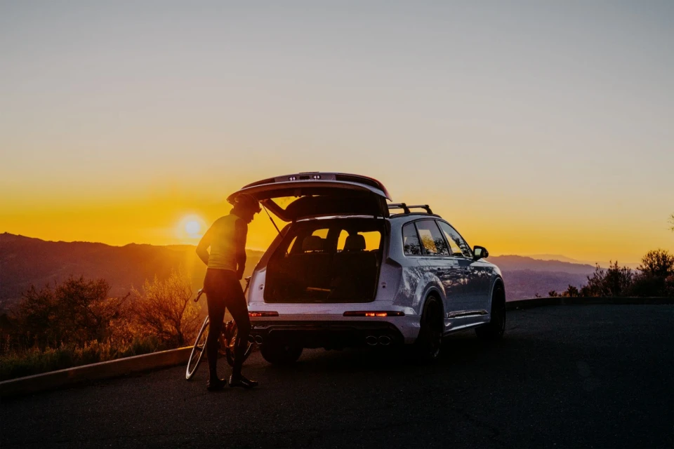 An adult cyclist next to a silver SUV with its trunk open on a road with a mountain and a bright sunset in the background.