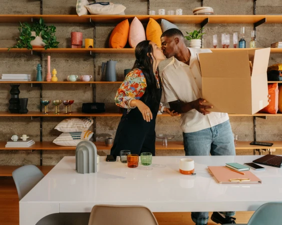 A young couple shares a kiss in their new home, surrounded by a white table and decorative shelves filled with colorful items.