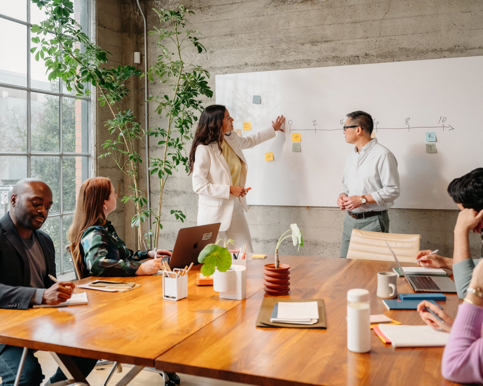 Diverse team collaborating in an office, with a woman presenting ideas on a whiteboard and colleagues engaged in discussion.