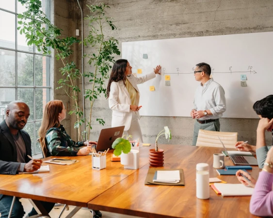 Diverse team collaborating in an office, with a woman presenting ideas on a whiteboard and colleagues engaged in discussion.