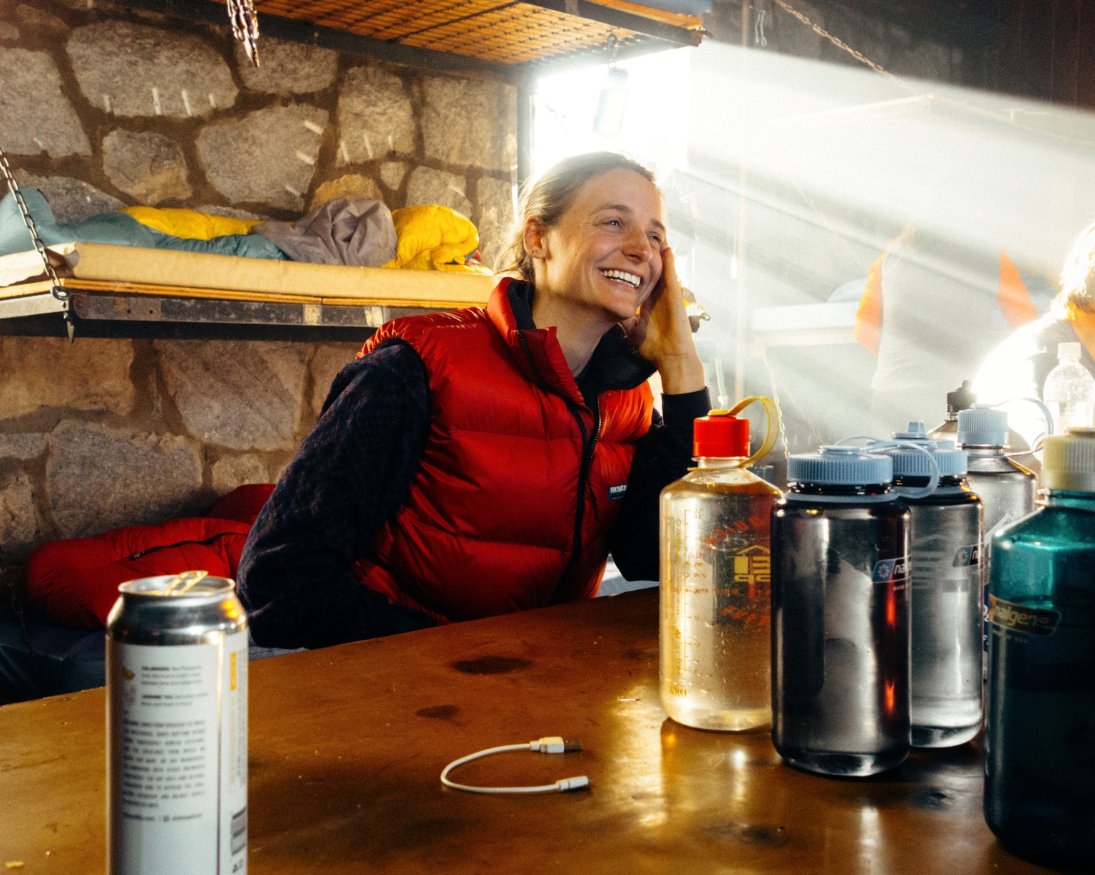 Smiling woman in a red vest sits at a table with water bottles in a stone shelter, lit by sunlight streaming through the doorway.