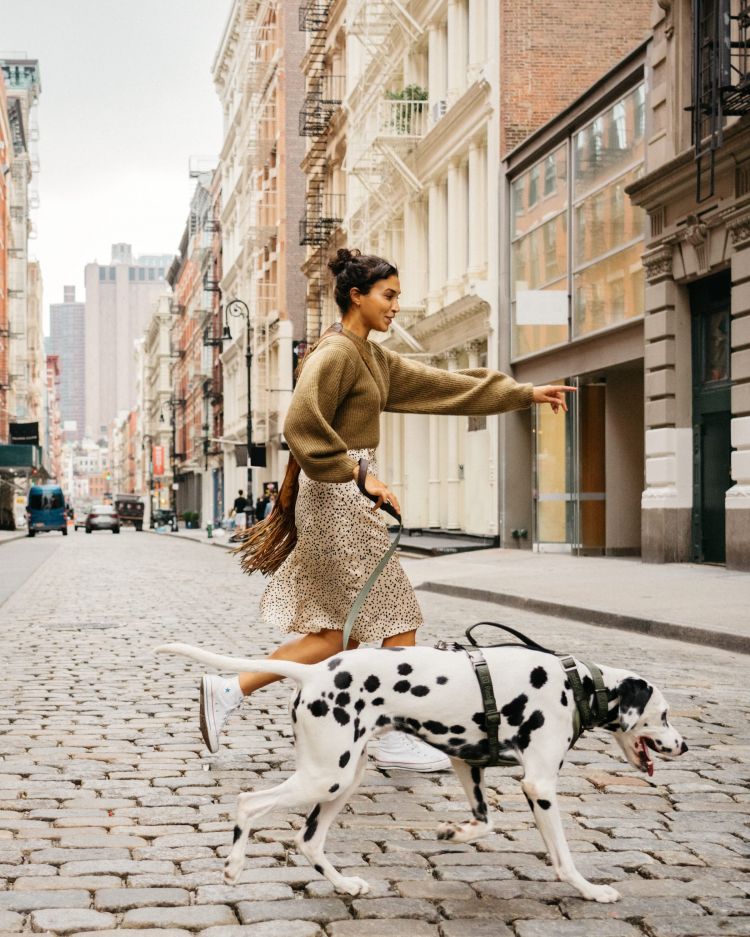 An adult female runs with a dalmatian dog on a city street, wearing a skirt and sweater with apartment buildings in the background.