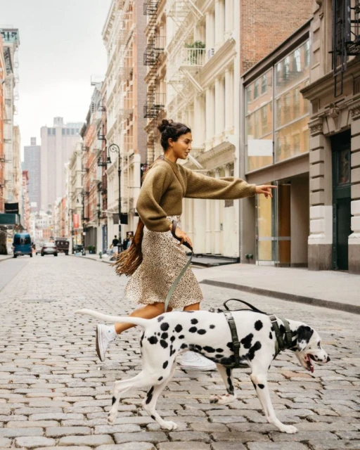 An adult female runs with a dalmatian dog on a city street, wearing a skirt and sweater with apartment buildings in the background.