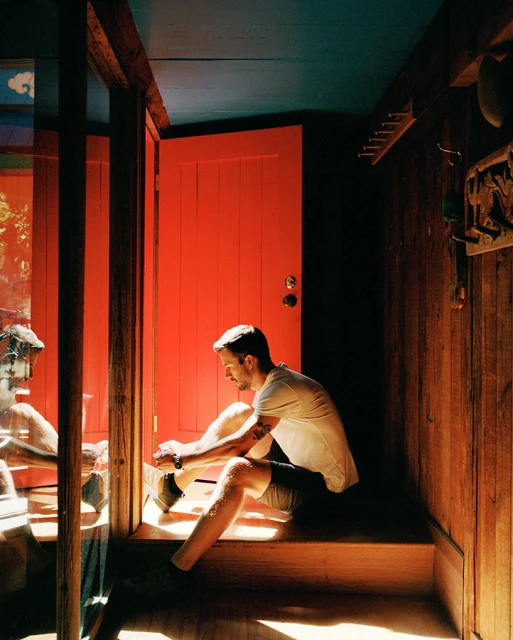 Man sitting on wooden stairs indoors, tying shoes in natural light. Red door visible in background, casual attire, focused expression.