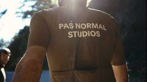 Man in cycling jersey with 'Pas Normal Studios' text, sunlit background. Sportswear, cyclist, outdoor apparel, fashion, athletic wear.