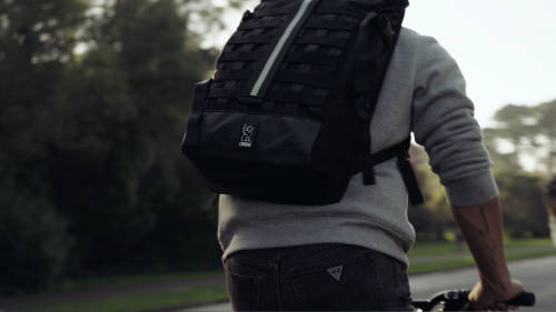 Person riding a bike on a path with a large black backpack, trees, and a bright sky in the background.