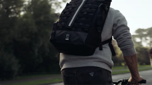 Person riding a bike on a path with a large black backpack, trees, and a bright sky in the background.