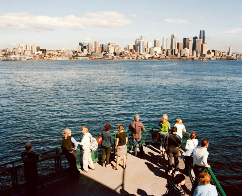 A group of people on a ferry look out at a city skyline across the water on a sunny day with blue sky and light clouds.