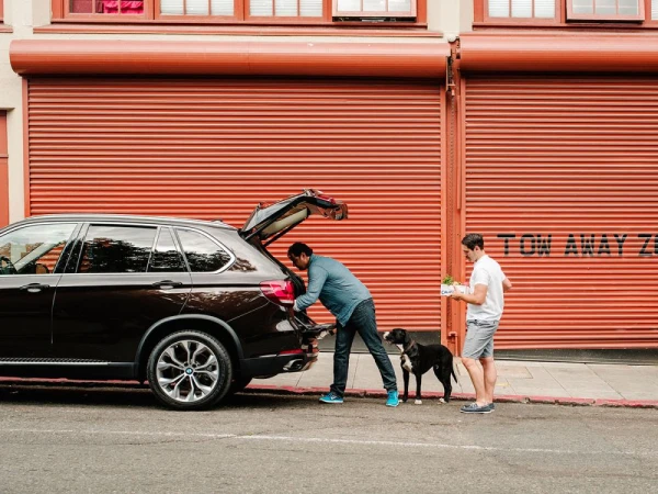 Brown SUV parked with two adult males and a black dog standing near a bright orange roll-up door in an urban setting.