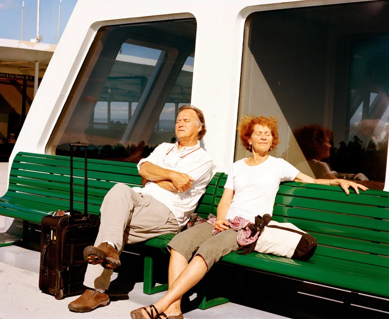 Older couple relaxing on ferry bench, bathed in warm sunlight, with luggage. Calm travel scene with relaxed vacationers.