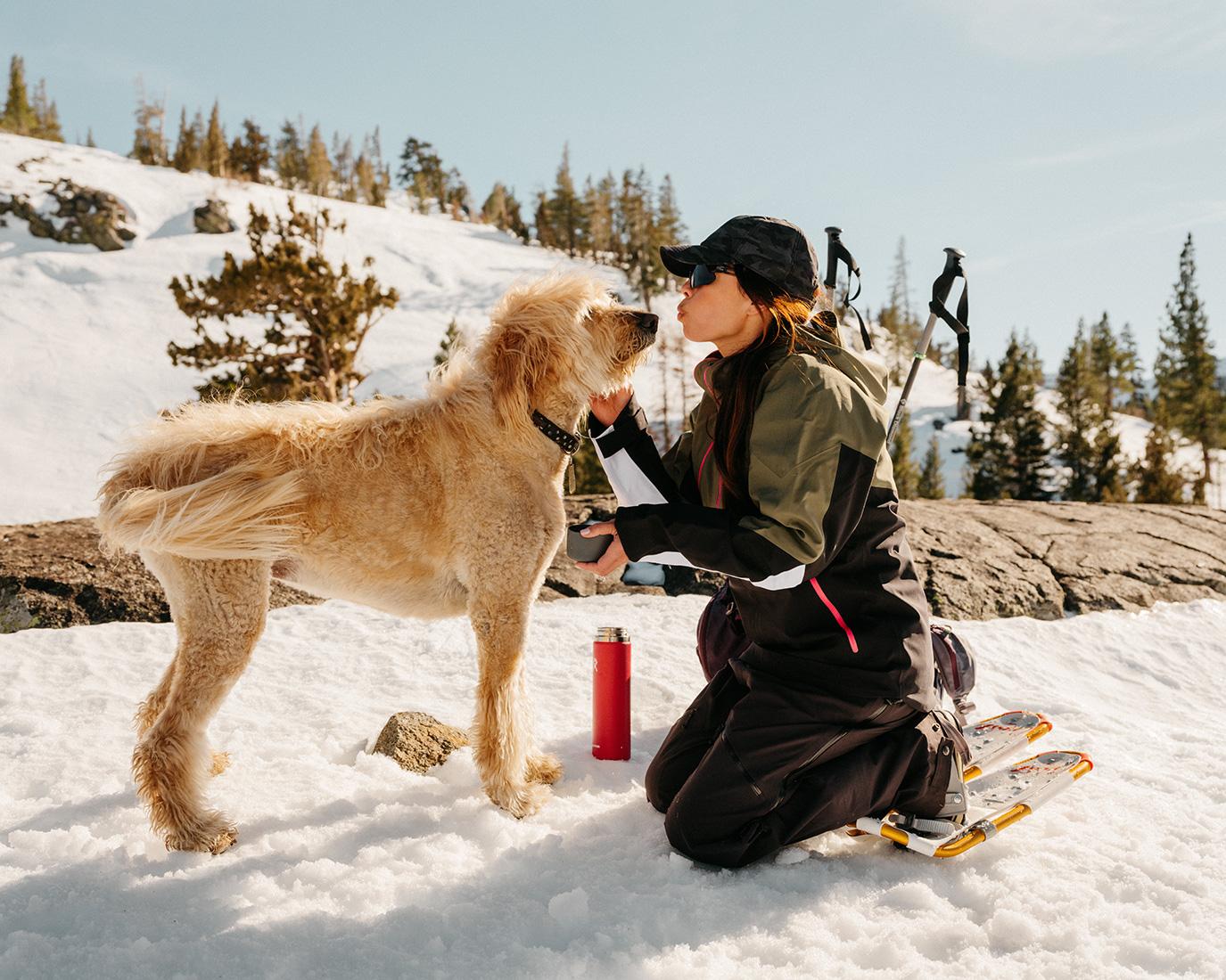 Woman outdoors in snow kneeling next to her dog, snowshoes and a red thermos visible, trees and a blue sky in the background.