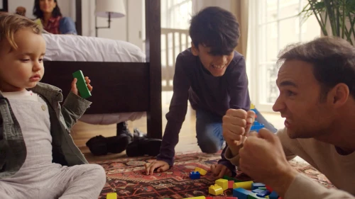 Father, son, and toddler playing with colorful toy blocks on a patterned rug in a brightly lit home; mother reads on bed in the background.