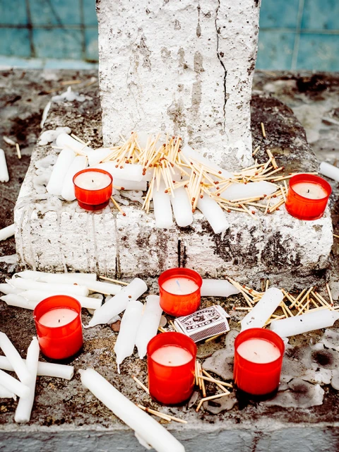 Several lit candles in red containers and unlit white candles and matches are on a weathered stone surface, creating a somber scene.