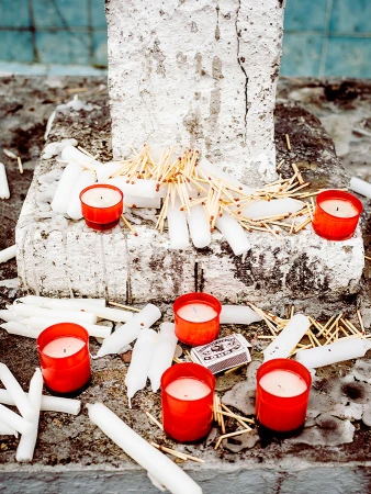 Several lit candles in red containers and unlit white candles and matches are on a weathered stone surface, creating a somber scene.