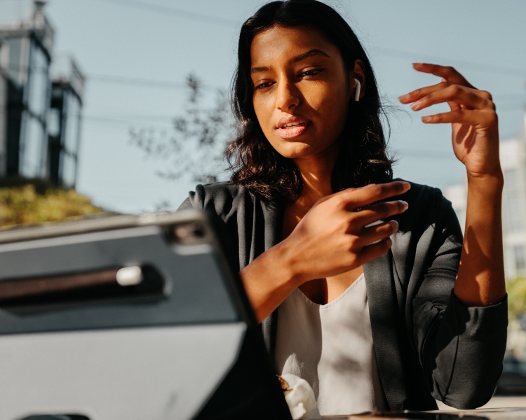 Adult female wearing a blazer and earpiece sits at a table using a tablet outdoors in sunlight with buildings and greenery in the background.