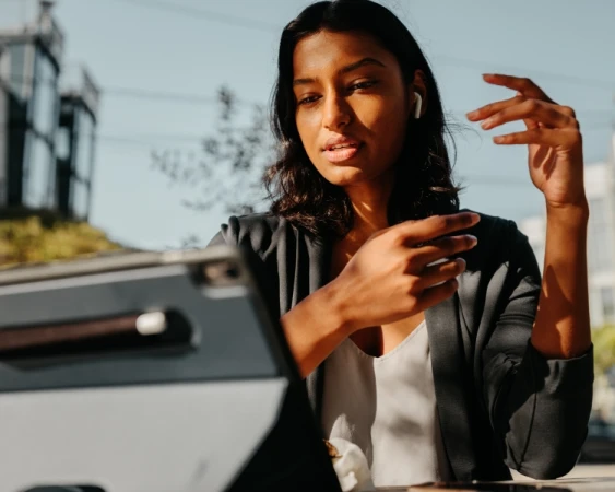 Adult female wearing a blazer and earpiece sits at a table using a tablet outdoors in sunlight with buildings and greenery in the background.