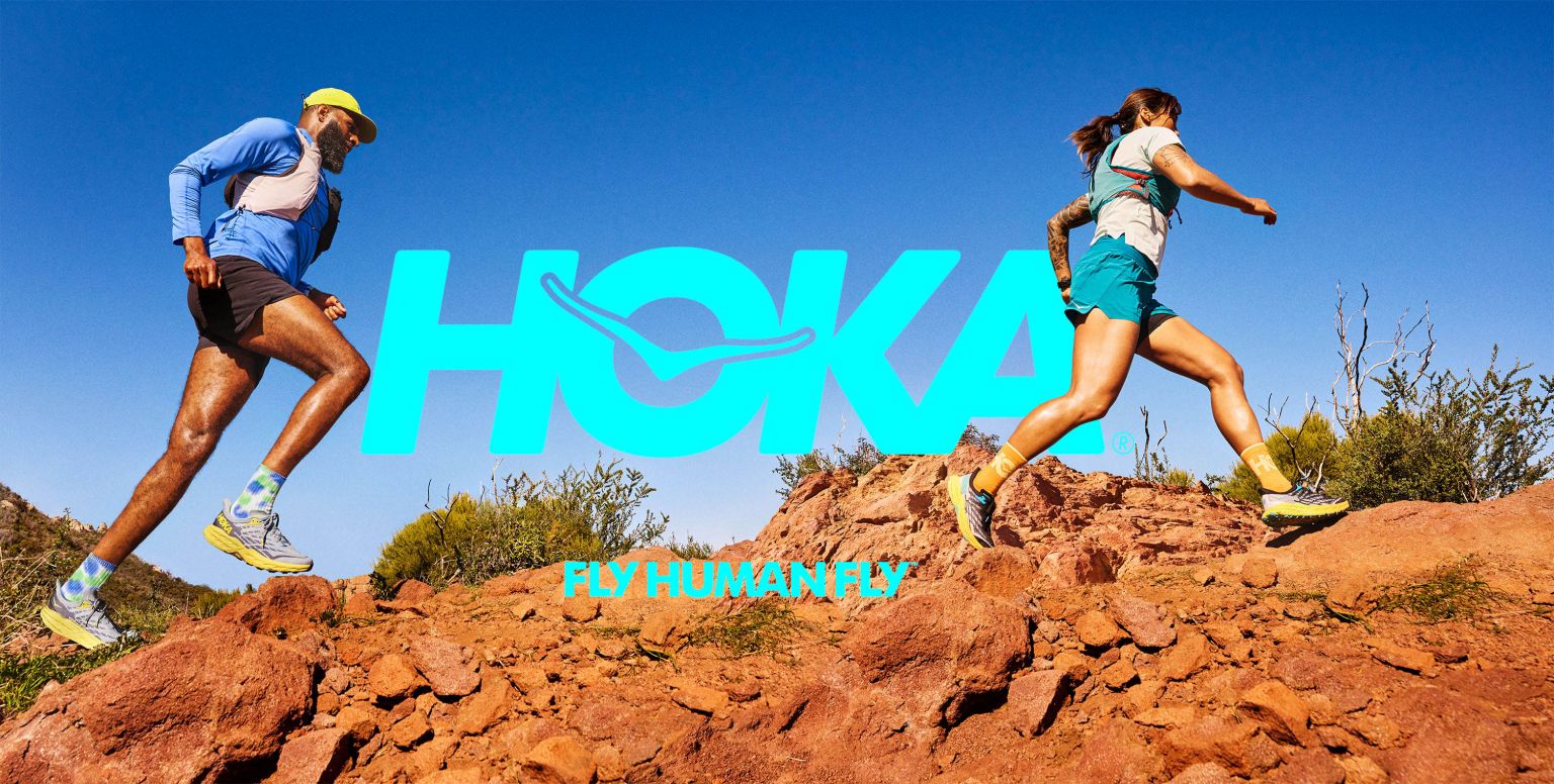 Two athletes, a male and female, run on a red rock trail against a blue sky. Hoka brand logo and slogan are overlaid.
