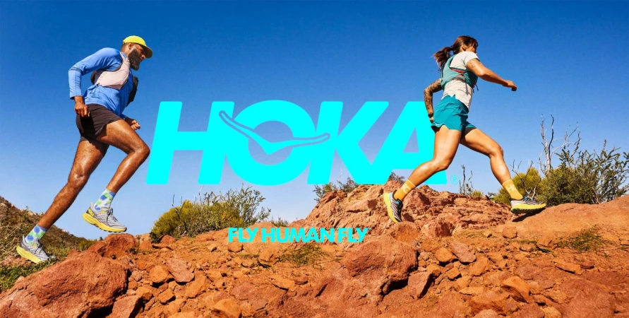 Two athletes, a male and female, run on a red rock trail against a blue sky. Hoka brand logo and slogan are overlaid.