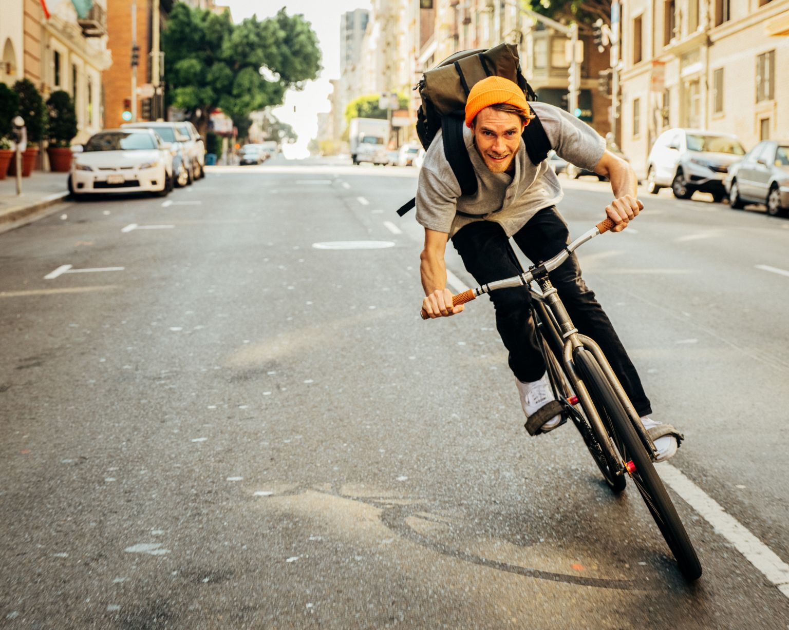 A cyclist wearing an orange beanie and backpack rides a bicycle down a city street on a sunny day, with cars and buildings blurred in the background.