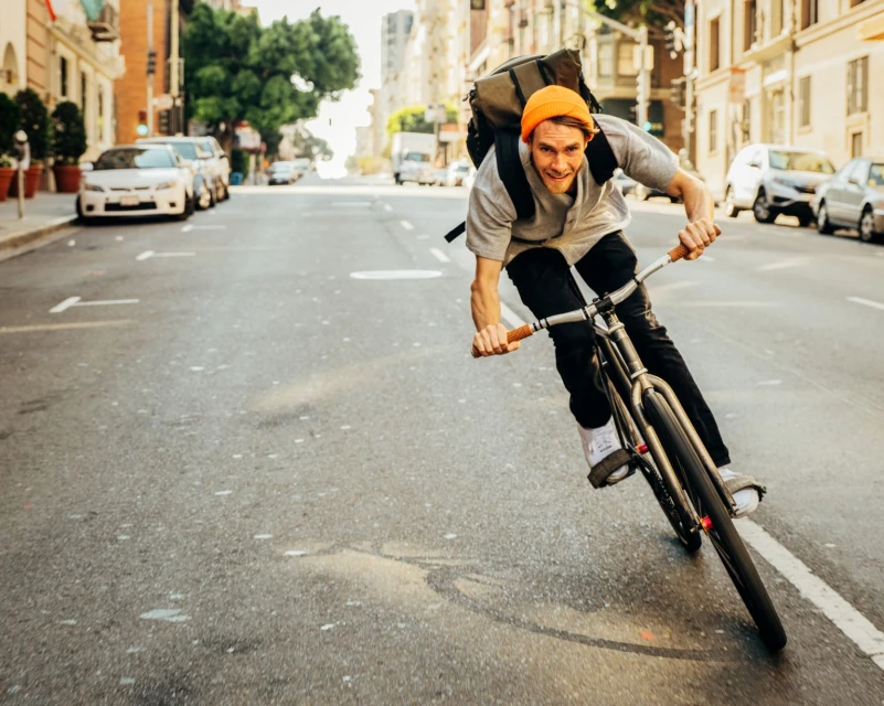 A cyclist wearing an orange beanie and backpack rides a bicycle down a city street on a sunny day, with cars and buildings blurred in the background.