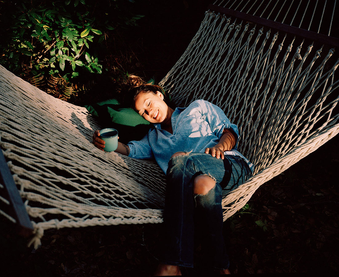 Woman relaxing in hammock with drink outdoors in the sun. She is lying down, wearing jeans and a denim shirt, with green foliage behind.