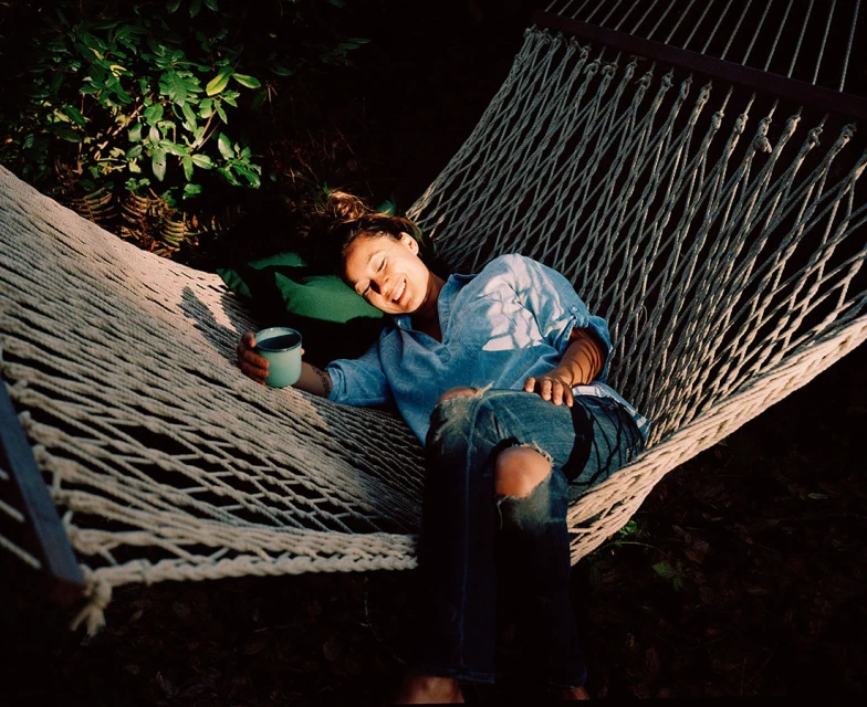 Woman relaxing in hammock with drink outdoors in the sun. She is lying down, wearing jeans and a denim shirt, with green foliage behind.