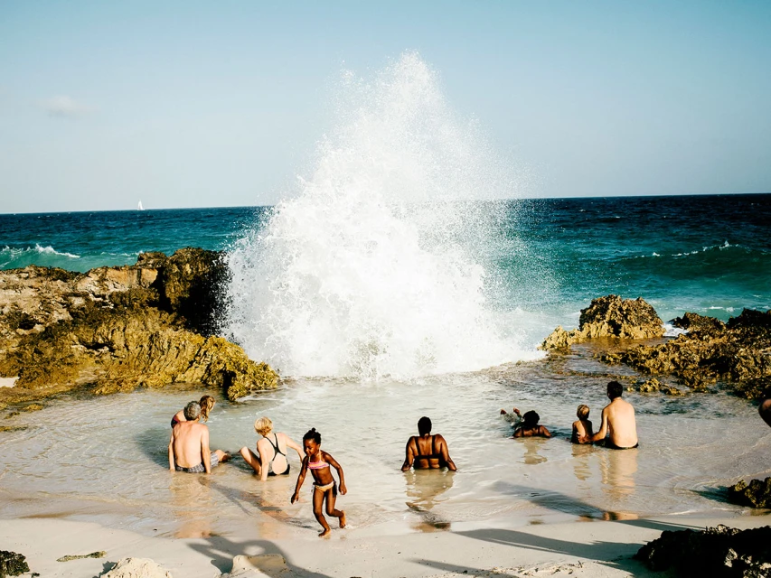 Ocean wave crashes near people relaxing on the sandy beach with rocky outcroppings; summer scene with turquoise water under a clear sky.