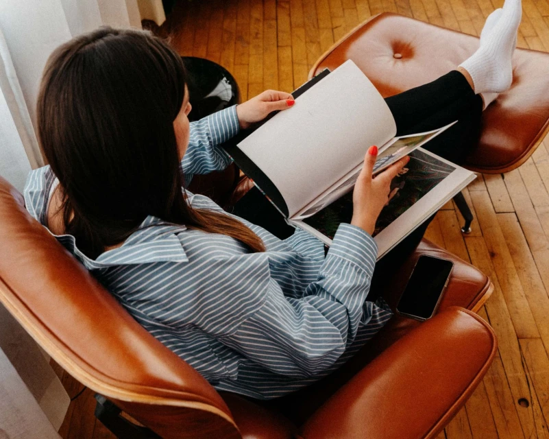 A woman sits in a leather chair with her feet up, reading a magazine in a relaxed pose in an indoor setting.