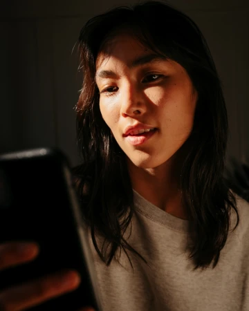 Indoor shot of a smiling Asian woman in natural light using a smartphone, wearing a grey t-shirt with a dark background.